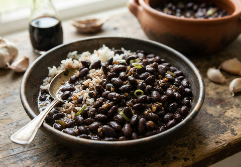 A rustic ceramic bowl filled with black beans over white rice, garnished with chopped green onions, set on a wooden table with garlic cloves, a small bottle, and another bowl in the background.